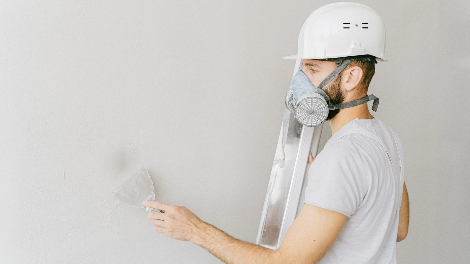 Vakman met helm en stofmasker werkt een binnenwand bij met een plamuurmes